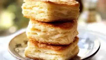 Three stacked golden brown flaky biscuits on a ceramic plate, with visible buttery layers and a soft, fluffy texture.