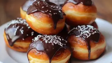 Close-up of golden Boston Cream Doughnut Holes stacked on a white plate, filled with pastry cream and topped with thick chocolate glaze and white sprinkles.