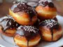Close-up of golden Boston Cream Doughnut Holes stacked on a white plate, filled with pastry cream and topped with thick chocolate glaze and white sprinkles.