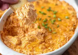 Close-up of creamy Buffalo Chicken Dip in a white baking dish, topped with melted cheddar cheese and green onions, with a cracker scooping out a cheesy portion.