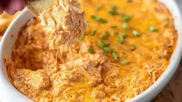 Close-up of creamy Buffalo Chicken Dip in a white baking dish, topped with melted cheddar cheese and green onions, with a cracker scooping out a cheesy portion.