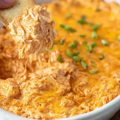 Close-up of creamy Buffalo Chicken Dip in a white baking dish, topped with melted cheddar cheese and green onions, with a cracker scooping out a cheesy portion.