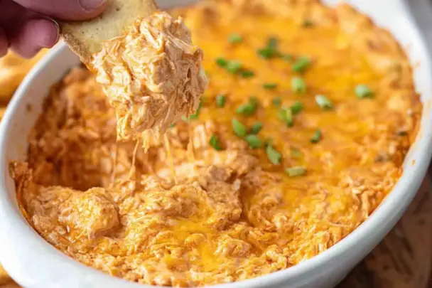Close-up of creamy Buffalo Chicken Dip in a white baking dish, topped with melted cheddar cheese and green onions, with a cracker scooping out a cheesy portion.