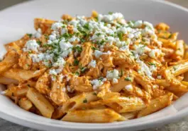 A close-up of creamy Buffalo Chicken Pasta made with penne, shredded chicken, spicy Buffalo sauce, and topped with crumbled cheese and chopped parsley in a white bowl.