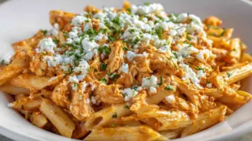 A close-up of creamy Buffalo Chicken Pasta made with penne, shredded chicken, spicy Buffalo sauce, and topped with crumbled cheese and chopped parsley in a white bowl.