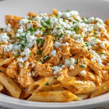 A close-up of creamy Buffalo Chicken Pasta made with penne, shredded chicken, spicy Buffalo sauce, and topped with crumbled cheese and chopped parsley in a white bowl.