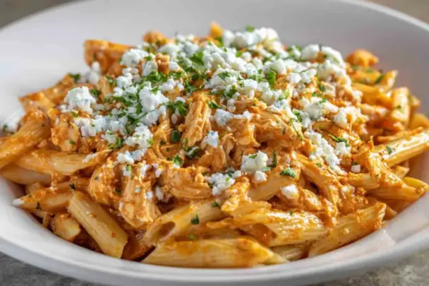 A close-up of creamy Buffalo Chicken Pasta made with penne, shredded chicken, spicy Buffalo sauce, and topped with crumbled cheese and chopped parsley in a white bowl.