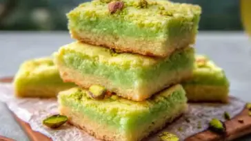 Stack of vibrant green and golden-brown pistachio sugar cookie bars on a wooden board lined with parchment, topped with chopped pistachios and surrounded by scattered nuts—showcasing their soft texture and layered look.