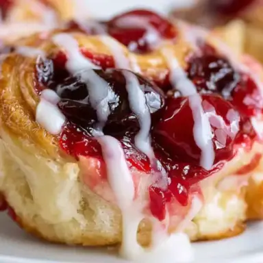 Close-up of a golden-brown cherry cheesecake pinwheel topped with cherry pie filling and drizzled icing, showing its flaky layers and creamy center.