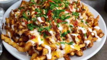A white plate filled with crispy crinkle-cut fries topped with seasoned chicken, melted cheese, ranch drizzle, diced tomatoes, and chopped parsley.