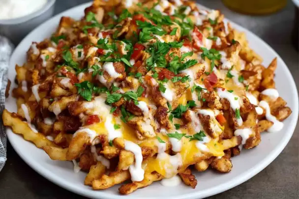A white plate filled with crispy crinkle-cut fries topped with seasoned chicken, melted cheese, ranch drizzle, diced tomatoes, and chopped parsley.