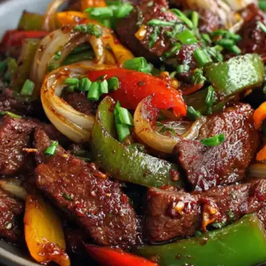 Close-up of Chinese Pepper Steak with sliced beef, red and green bell peppers, onions, and thick pepper sauce, garnished with chopped green onions on a dark plate.