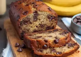 Sliced chocolate chip banana bread loaf on a wooden board, showing a moist, golden interior loaded with melted chocolate chips, ripe bananas in the background