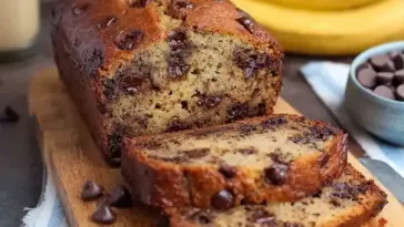 Sliced chocolate chip banana bread loaf on a wooden board, showing a moist, golden interior loaded with melted chocolate chips, ripe bananas in the background