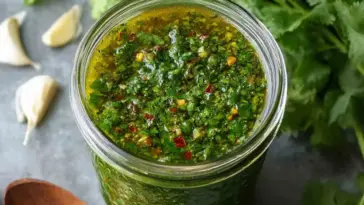 Close-up of a glass jar filled with vibrant green cilantro chimichurri sauce, surrounded by garlic cloves and fresh cilantro leaves on a gray background.