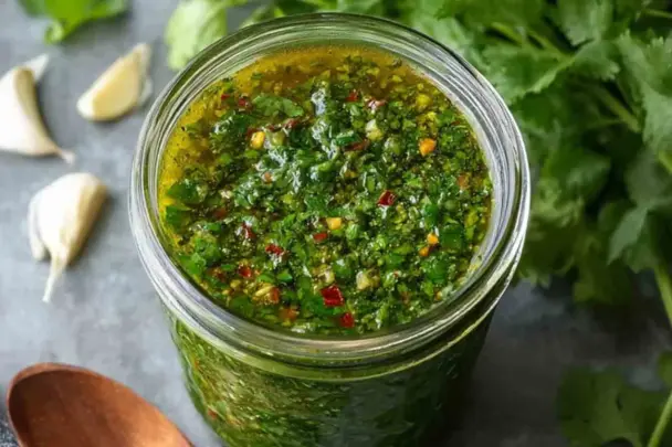 Close-up of a glass jar filled with vibrant green cilantro chimichurri sauce, surrounded by garlic cloves and fresh cilantro leaves on a gray background.