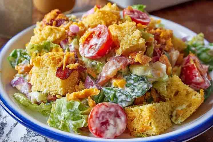 Close-up of Southern cornbread salad featuring crumbled cornbread, cherry tomatoes, bacon, and lettuce, all mixed with creamy dressing and served in a white enamel bowl.