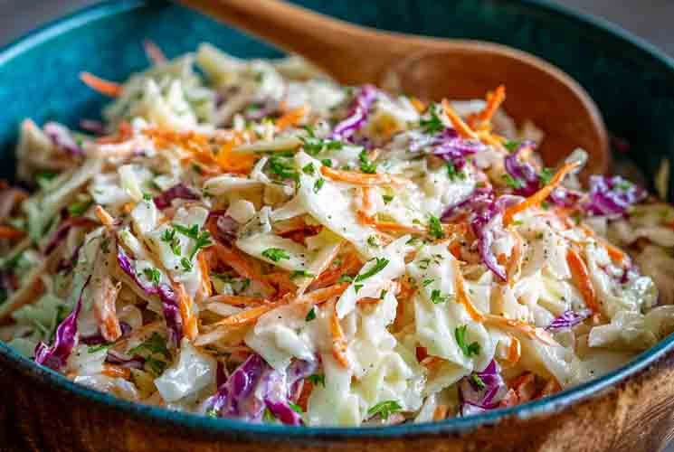 Overhead view of a vibrant creamy cole slaw with green and purple cabbage, shredded carrots, and fresh parsley in a blue serving bowl with a wooden spoon.
