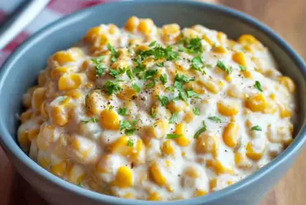 Close-up of a bowl filled with slow cooker creamed corn, topped with black pepper and fresh parsley, in a blue ceramic bowl on a wooden table.