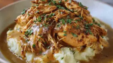 Plate of shredded crockpot chicken in rich brown gravy served over mashed potatoes and garnished with chopped fresh parsley.