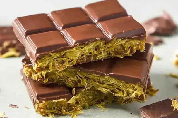 Close-up of stacked homemade Dubai chocolate bars filled with creamy pistachio and crispy golden kataifi pastry, coated in glossy dark chocolate.