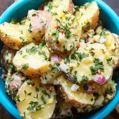 A vibrant bowl of French Potato Salad made with halved red potatoes, finely chopped parsley, red onion, and Dijon vinaigrette, served in a bright blue dish on a rustic wooden table.