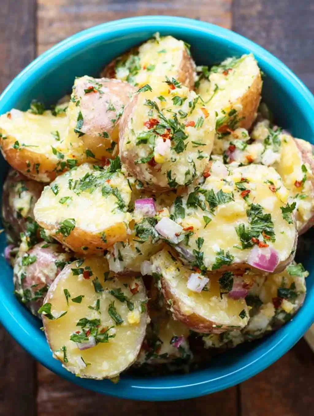 A vibrant bowl of French Potato Salad made with halved red potatoes, finely chopped parsley, red onion, and Dijon vinaigrette, served in a bright blue dish on a rustic wooden table.