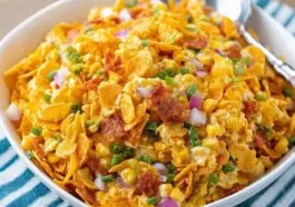 Vibrant bowl of Frito Corn Salad with golden corn chips, red onion, corn, and green onions in a creamy dressing, served with a silver spoon.