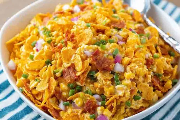 Vibrant bowl of Frito Corn Salad with golden corn chips, red onion, corn, and green onions in a creamy dressing, served with a silver spoon.