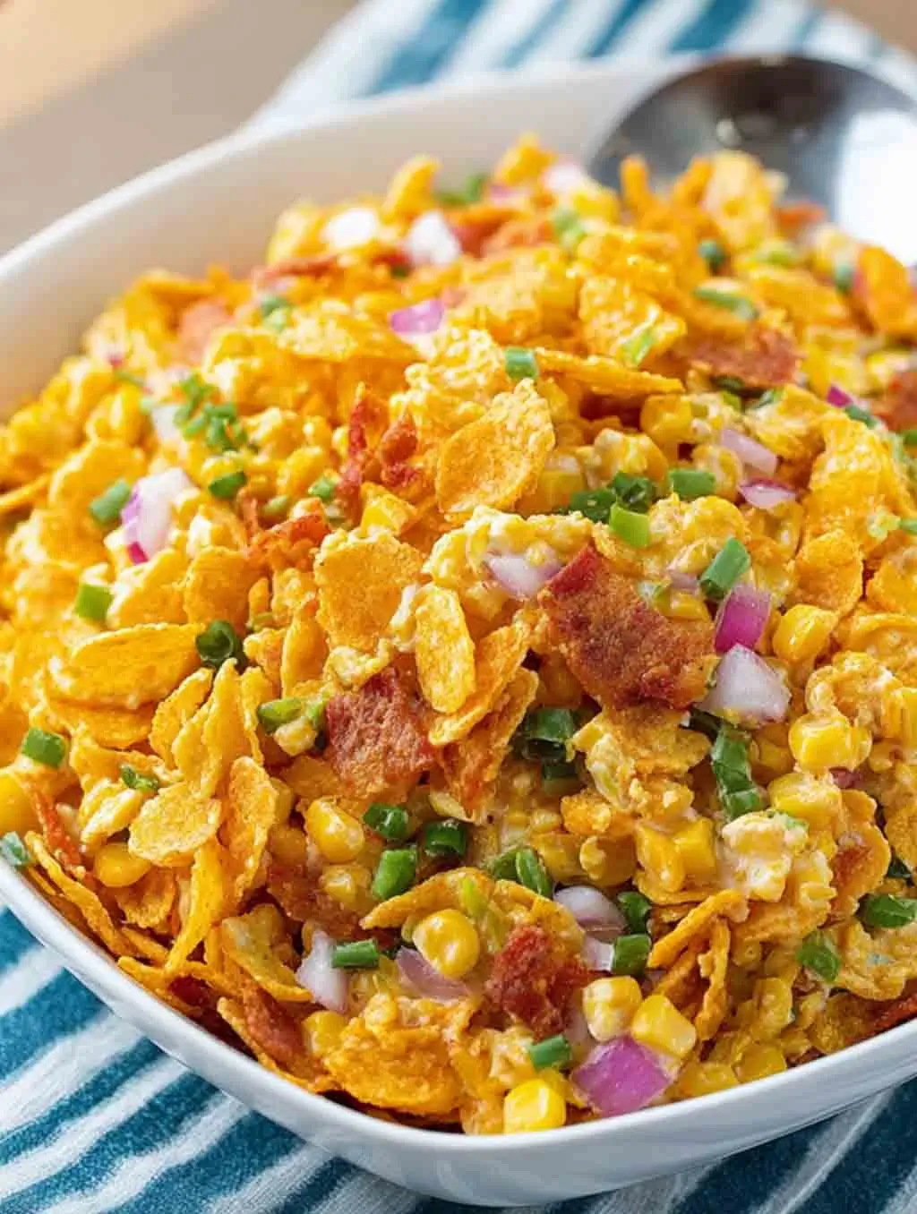 Close-up of a bowl of creamy Frito Corn Salad with corn, red onion, green peppers, and crushed Chili Cheese Fritos, served on a striped cloth.