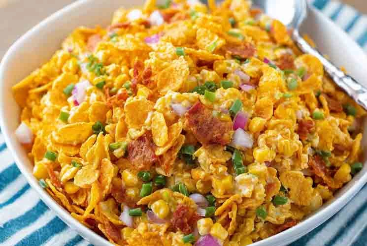 Vibrant bowl of Frito Corn Salad with golden corn chips, red onion, corn, and green onions in a creamy dressing, served with a silver spoon.