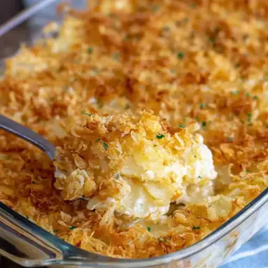 A glass baking dish filled with golden-brown, cheesy Funeral Potatoes with a crispy cornflake topping, with a serving spoon scooping out the creamy potato casserole.