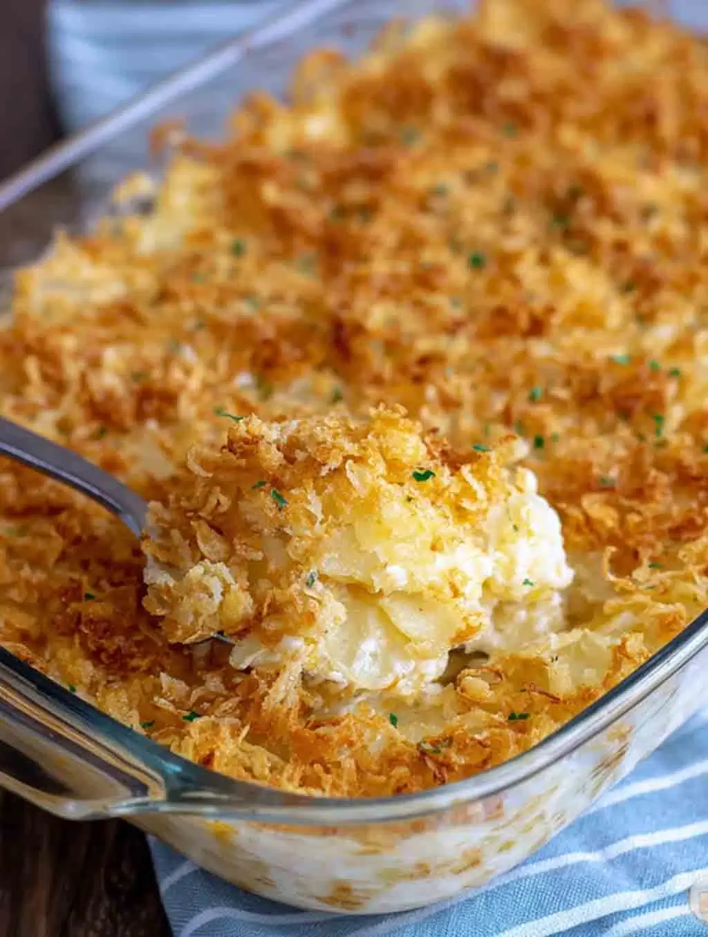 A glass baking dish filled with golden-brown, cheesy Funeral Potatoes with a crispy cornflake topping, with a serving spoon scooping out the creamy potato casserole.