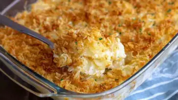 Golden and crispy funeral potatoes in a glass casserole dish, topped with toasted cornflakes and scooped to reveal a creamy, cheesy potato filling.