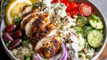 A colorful Greek chicken bowl with grilled marinated chicken, herbed rice, cucumber slices, cherry tomatoes, red onion, kalamata olives, feta cheese, and tzatziki sauce, garnished with lemon slices and fresh herbs.