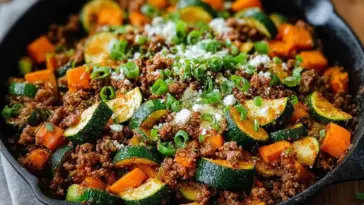 Close-up of a one-pan Ground Beef Zucchini Sweet Potato Skillet topped with green onions and cheese, served in a black cast iron skillet.
