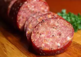 Close-up of sliced homemade summer sausage with visible spices and fat marbling, arranged on a wooden cutting board with parsley garnish in the background.