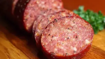Close-up of sliced homemade summer sausage with visible spices and fat marbling, arranged on a wooden cutting board with parsley garnish in the background.
