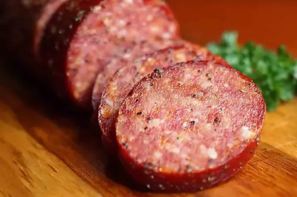 Close-up of sliced homemade summer sausage with visible spices and fat marbling, arranged on a wooden cutting board with parsley garnish in the background.