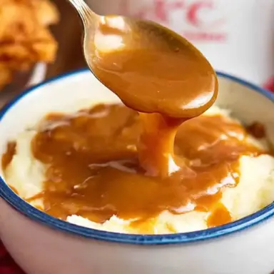 A spoon pouring rich, brown KFC-style gravy over a bowl of creamy mashed potatoes, with crispy fried chicken and a KFC container in the background.