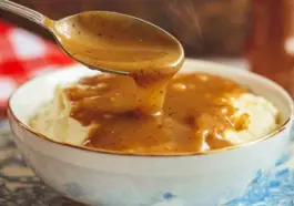A spoon drizzling hot, peppery brown KFC-style gravy over a bowl of creamy mashed potatoes in a white and blue bowl with steam rising.