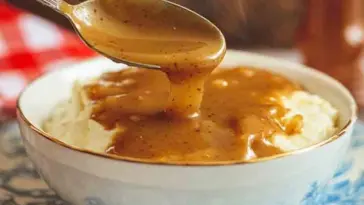 A spoon drizzling hot, peppery brown KFC-style gravy over a bowl of creamy mashed potatoes in a white and blue bowl with steam rising.