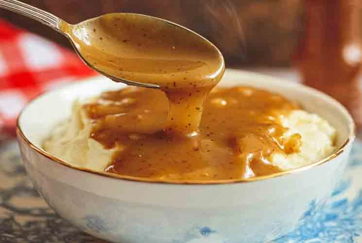 A spoon drizzling hot, peppery brown KFC-style gravy over a bowl of creamy mashed potatoes in a white and blue bowl with steam rising.