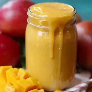 A thick, creamy mango smoothie overflowing from a glass jar, surrounded by ripe red mangoes and freshly diced mango chunks on a wooden surface.