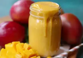 A thick, creamy mango smoothie overflowing from a glass jar, surrounded by ripe red mangoes and freshly diced mango chunks on a wooden surface.