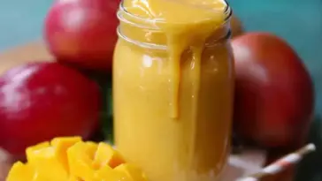 A thick, creamy mango smoothie overflowing from a glass jar, surrounded by ripe red mangoes and freshly diced mango chunks on a wooden surface.