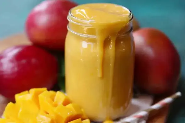 A thick, creamy mango smoothie overflowing from a glass jar, surrounded by ripe red mangoes and freshly diced mango chunks on a wooden surface.