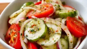 A close-up of a vibrant marinated salad featuring sliced cucumbers, tomatoes, and onions coated in a herb vinaigrette, served in a white bowl.