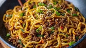 A close-up of Mongolian Ground Beef Noodles served in a bowl, topped with chopped green onions and sesame seeds, showcasing saucy noodles and crumbled beef in a rich brown glaze.
