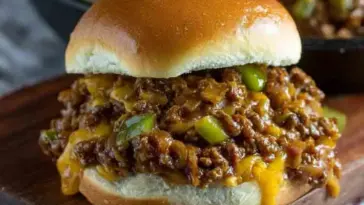 Juicy Philly Cheesesteak Sloppy Joe sandwich filled with cheesy ground beef, sautéed onions, and green peppers on a toasted bun, served on a wooden board.